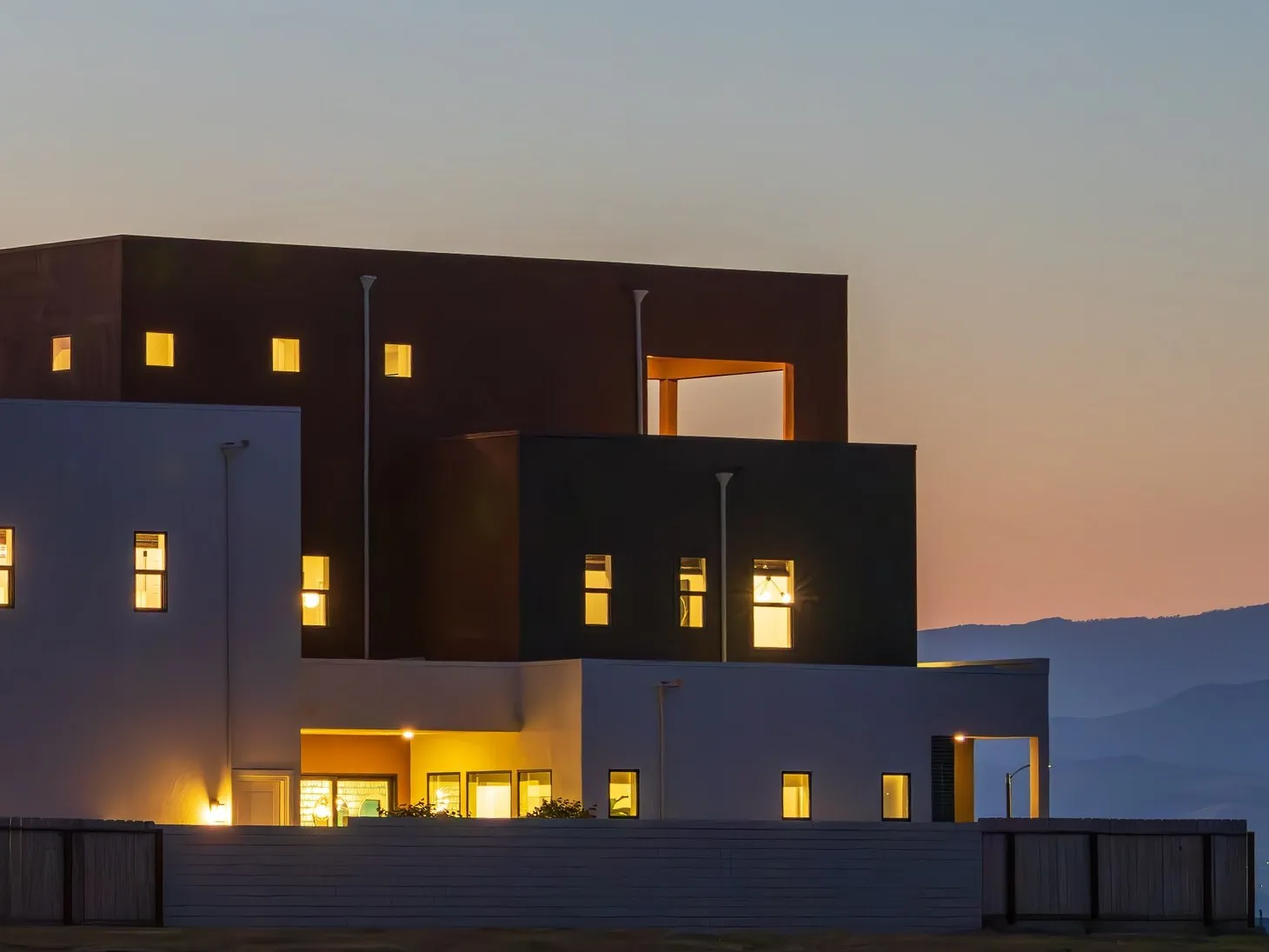 Modern cubic house facade in white and rust at dusk, lit windows